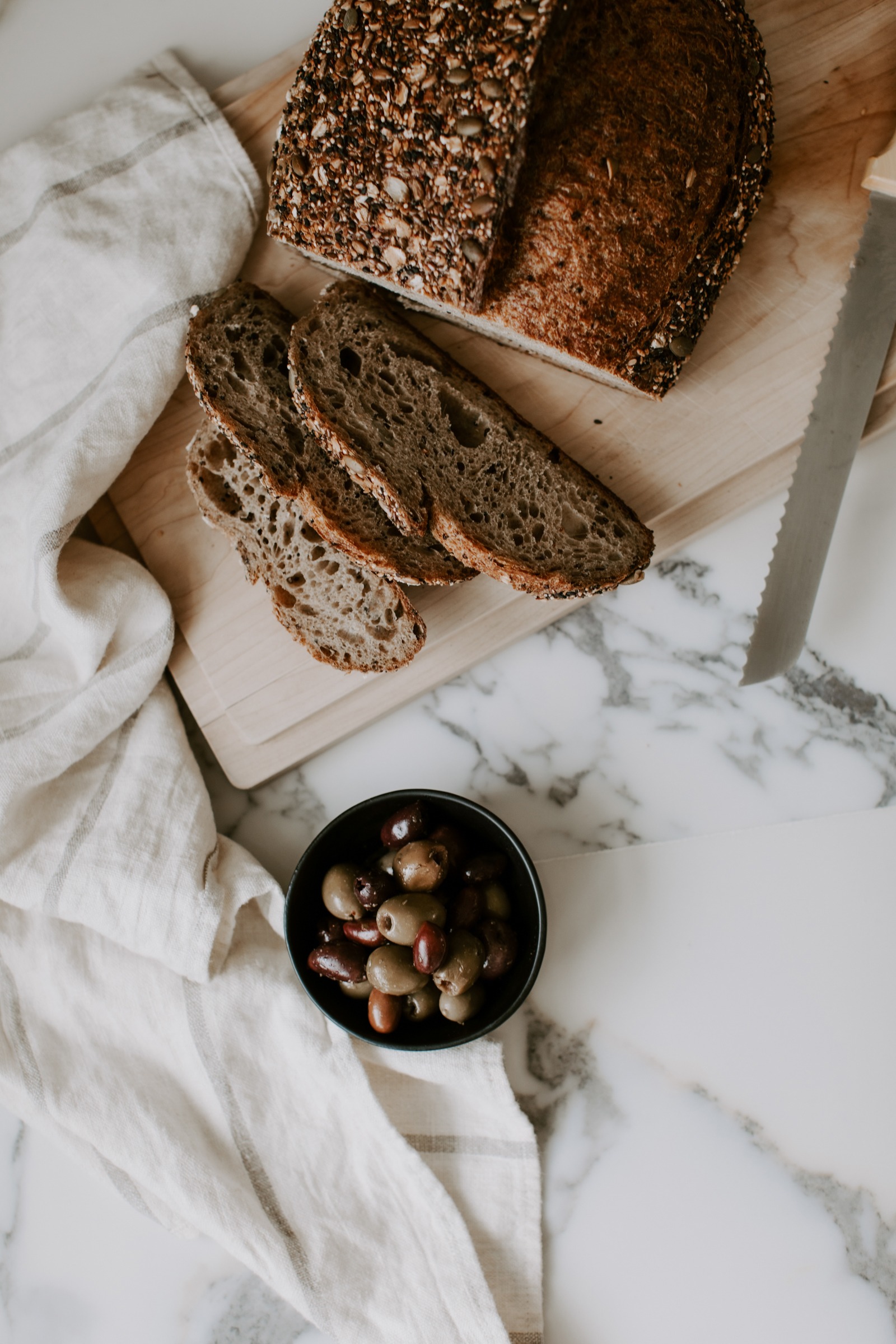Fresh artisan bread sliced on a wooden cutting board with a bowl of mixed olives, a linen cloth, and a marble countertop.