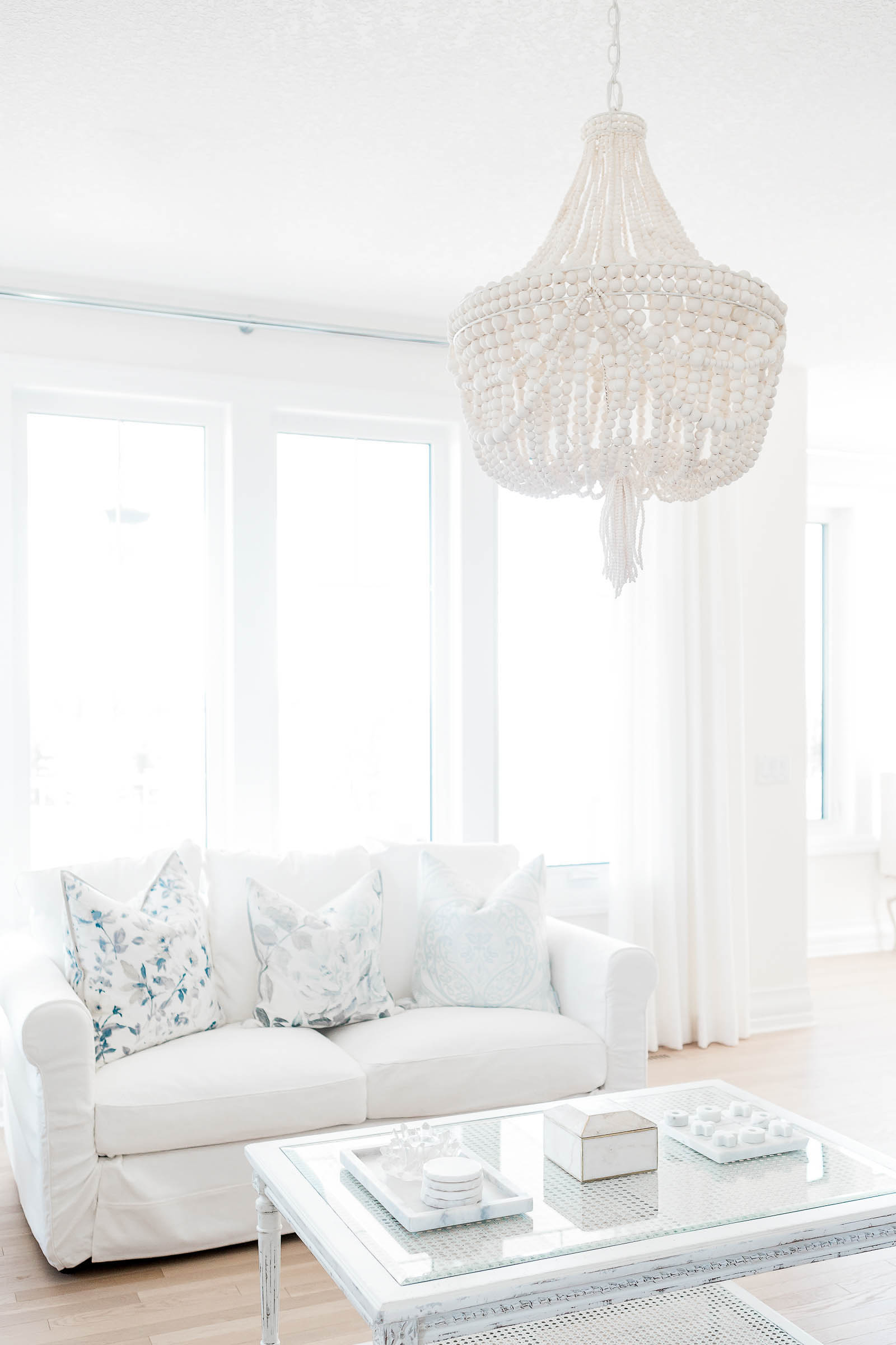 Elegant white living room with a crystal beaded chandelier, slipcovered sofa, decorative pillows, and a glass coffee table.