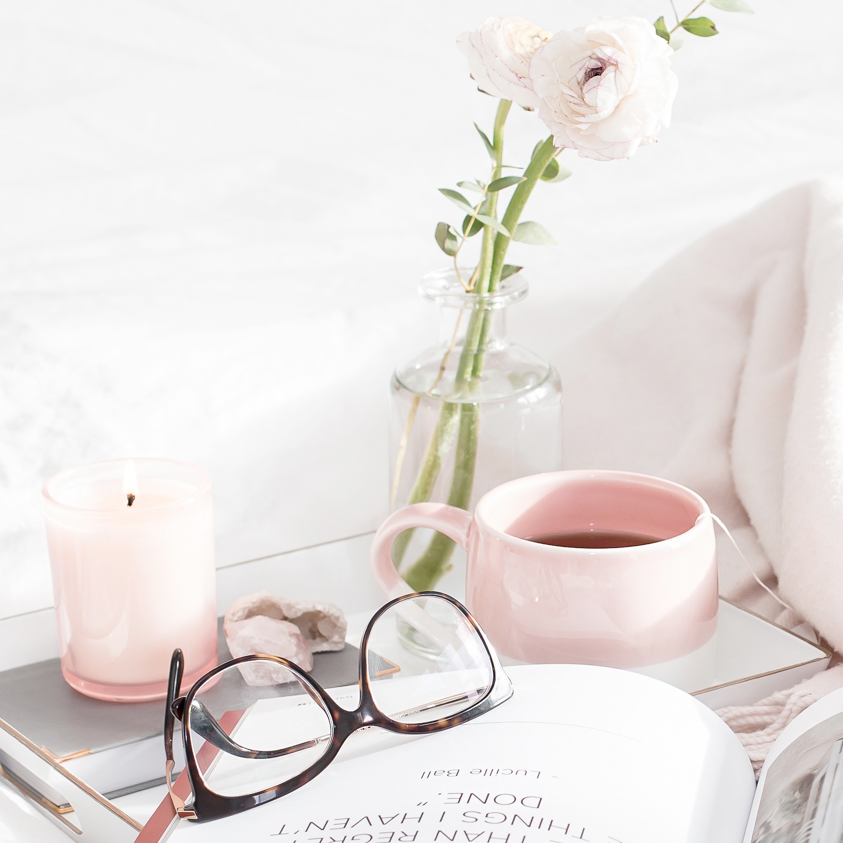 Open book beside a pink mug of tea, fresh flowers, a lit candle, and reading glasses on a bedside table.