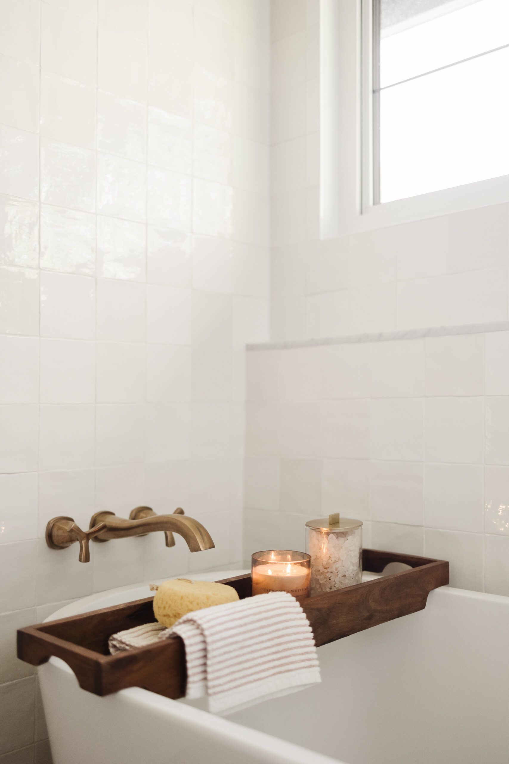 Freestanding bathtub with a wooden bath tray holding a candle, bath salts, natural sponge, and striped hand towel.