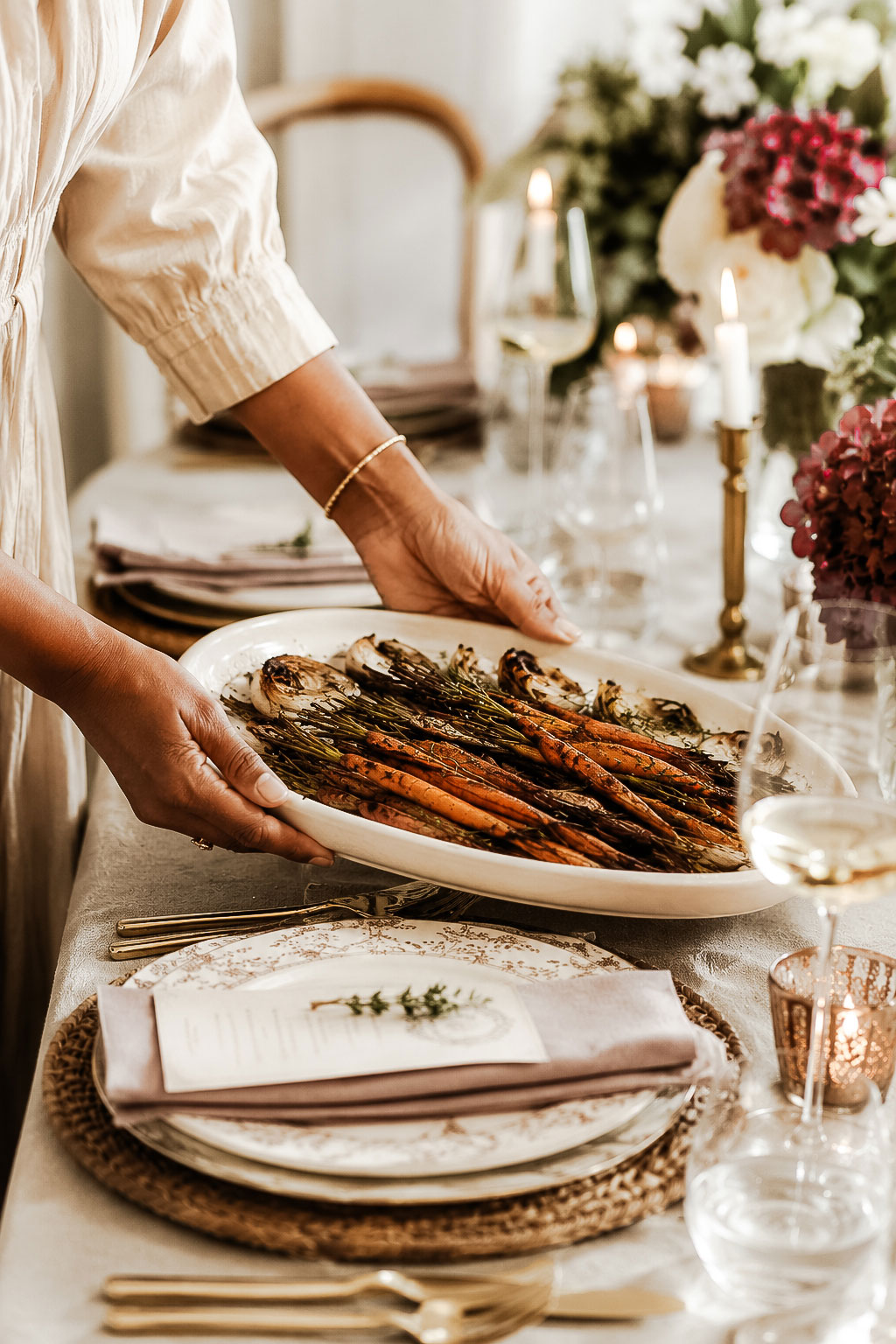 Hands placing a platter of roasted vegetables onto an elegantly set dining table with candles, flowers, and linen place settings.