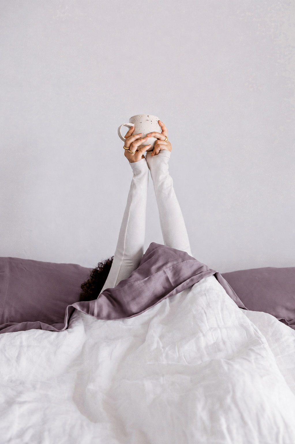 Hands holding a warm cup of coffee above crisp white bedding with soft plum linens during a slow, peaceful morning at home.